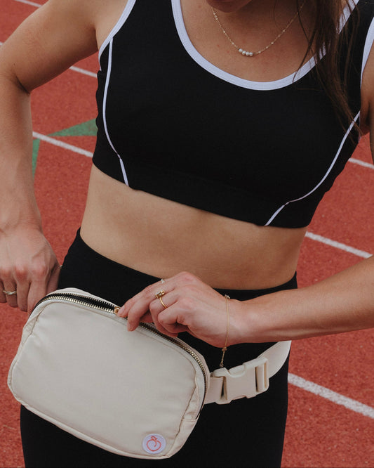 A woman on a running track wearing a black sports bra and black leggings, holding a cream-colored waist bag with a white strap.