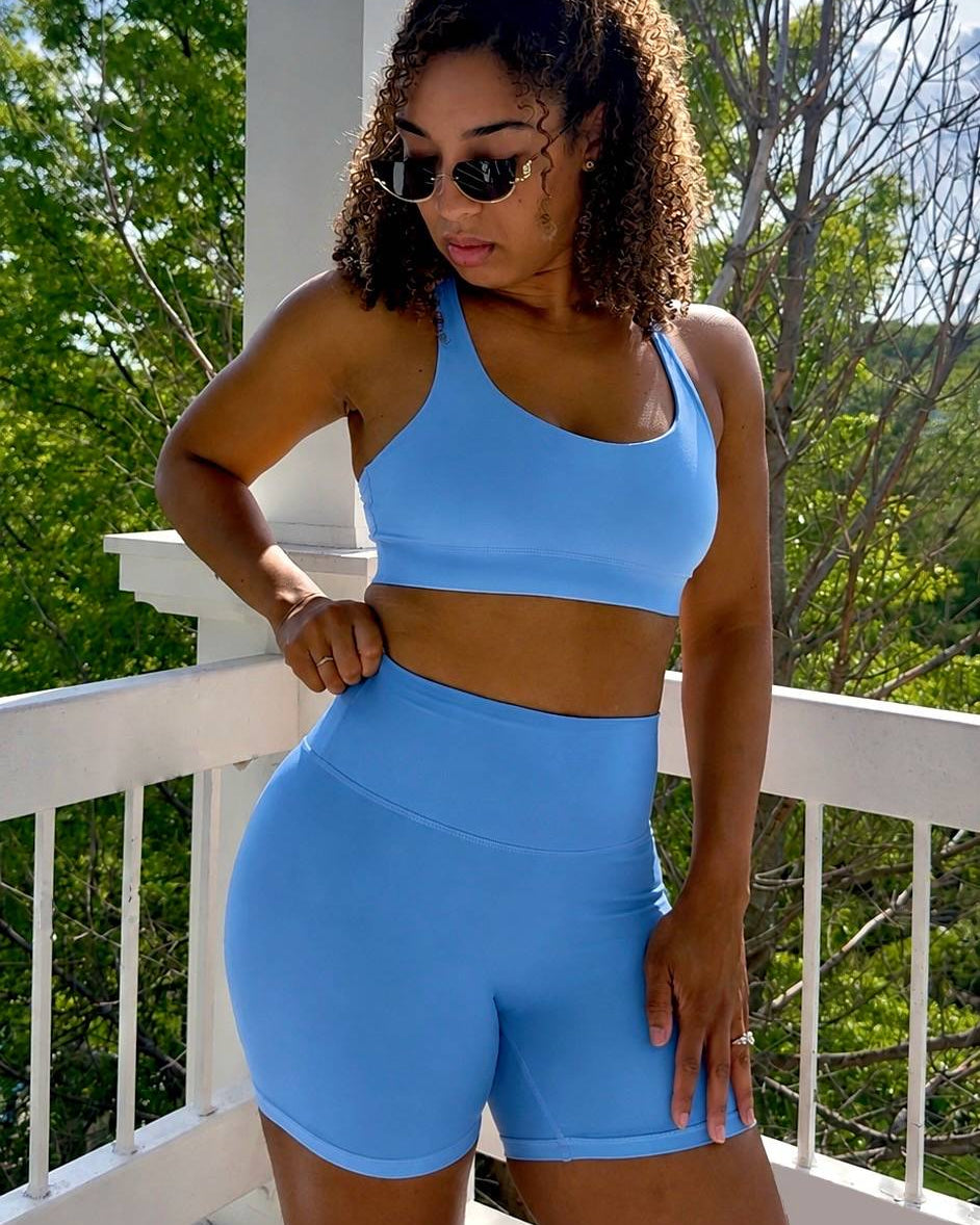 Woman wearing a blue sports bra and shorts set on a balcony with green trees in the background