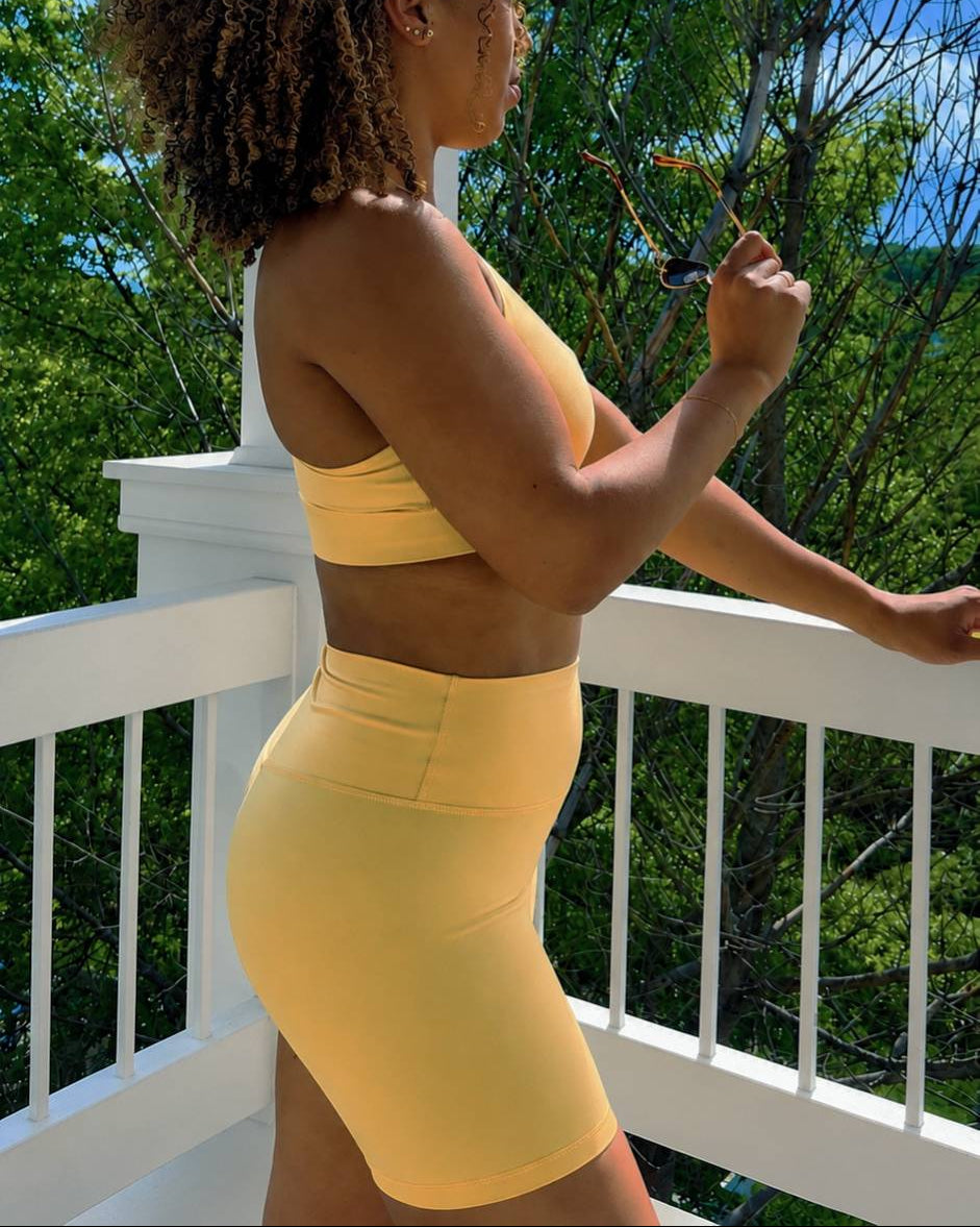 Woman in yellow athletic wear standing on a balcony with green trees and blue sky in the background