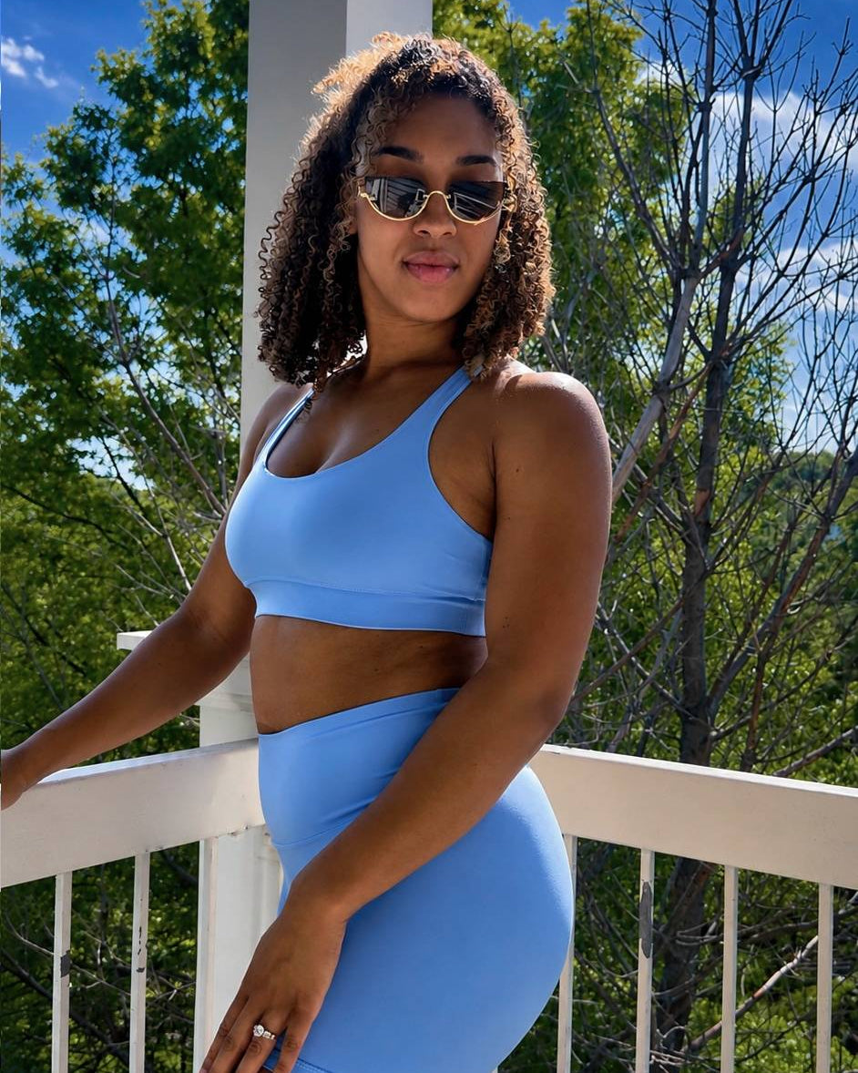 Woman in a blue sports bra and shorts sitting on a railing with trees and blue sky in the background