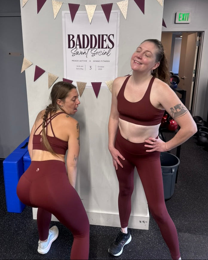 Two women in matching burgundy athletic wear posing in front of a gym wall with a 'Baddies Sweat Social' sign. One is standing and smiling, while the other is bent forward in a side stretch.