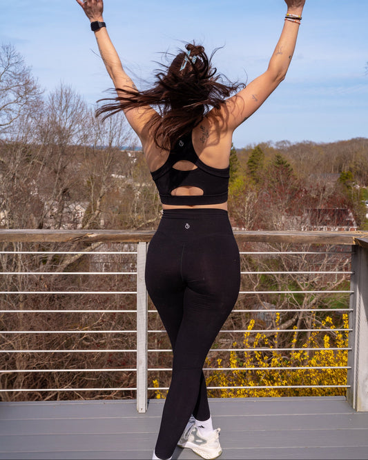 A woman viewed from behind wearing a black athletic sports bra with cutouts and matching black leggings, standing on an outdoor deck with her arms raised against a blue sky and trees.