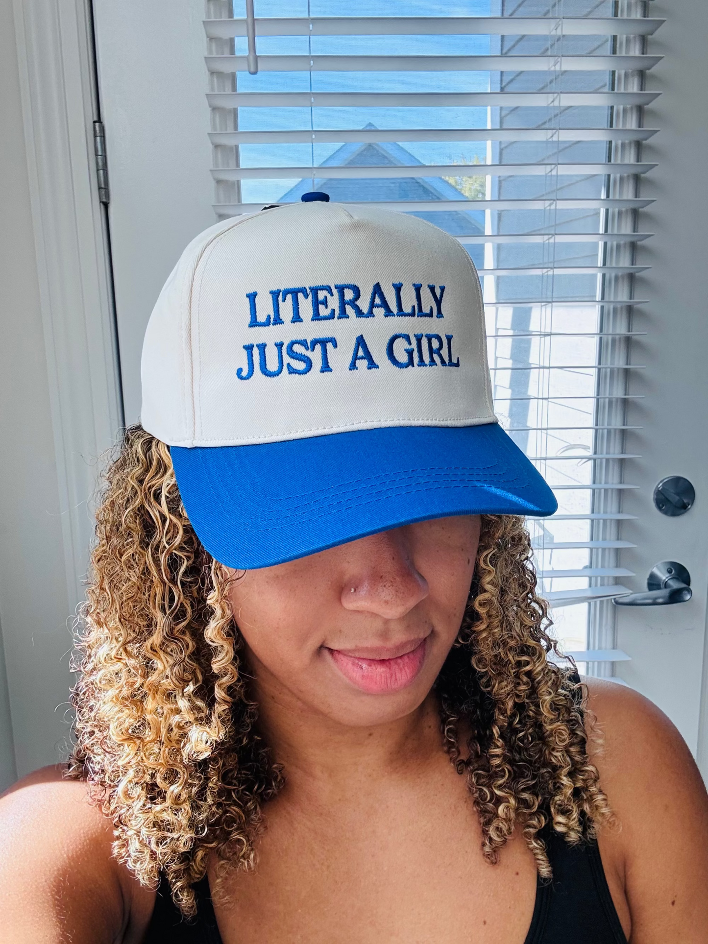 Person wearing a white and blue cap with 'LITERALLY JUST A GIRL' text in front of a window.