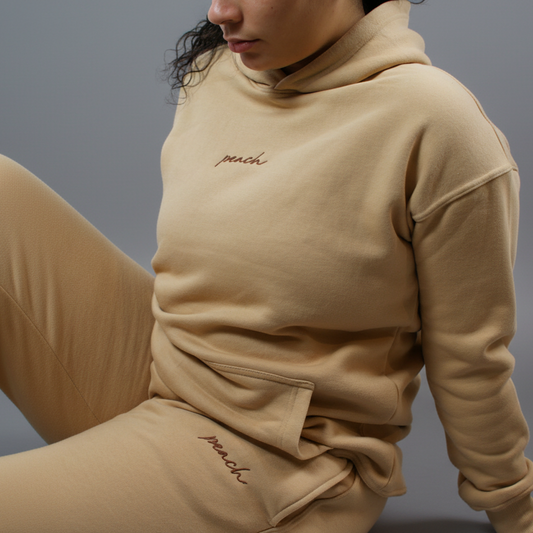 Person wearing a beige hoodie with 'peach' embroidery on a light background