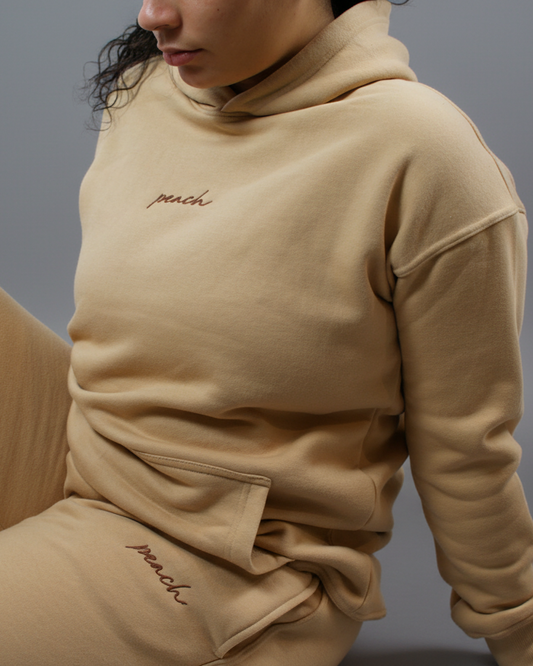 Person wearing a beige hoodie with 'peach' embroidery on a light background