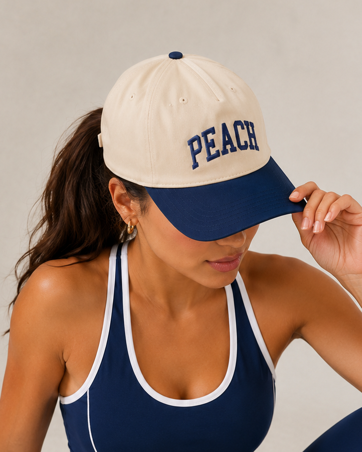 A woman wearing a navy blue and white athletic set consisting of a sports bra, leggings, and a matching baseball cap with 'PEACH' embroidered on the front, posing against a neutral studio backdrop.