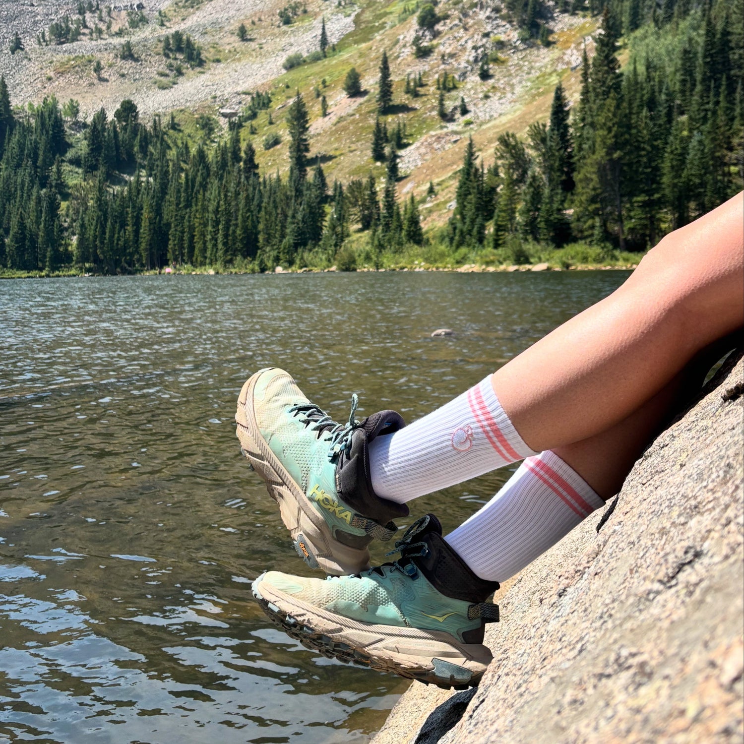 Person wearing hiking boots and socks sitting on a rock by a lake with mountains in the background