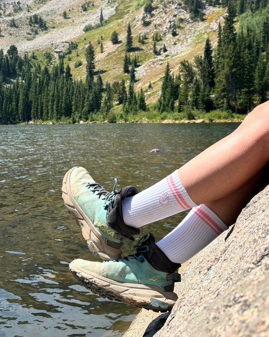 Person wearing hiking boots and socks sitting on a rock by a lake with mountains in the background