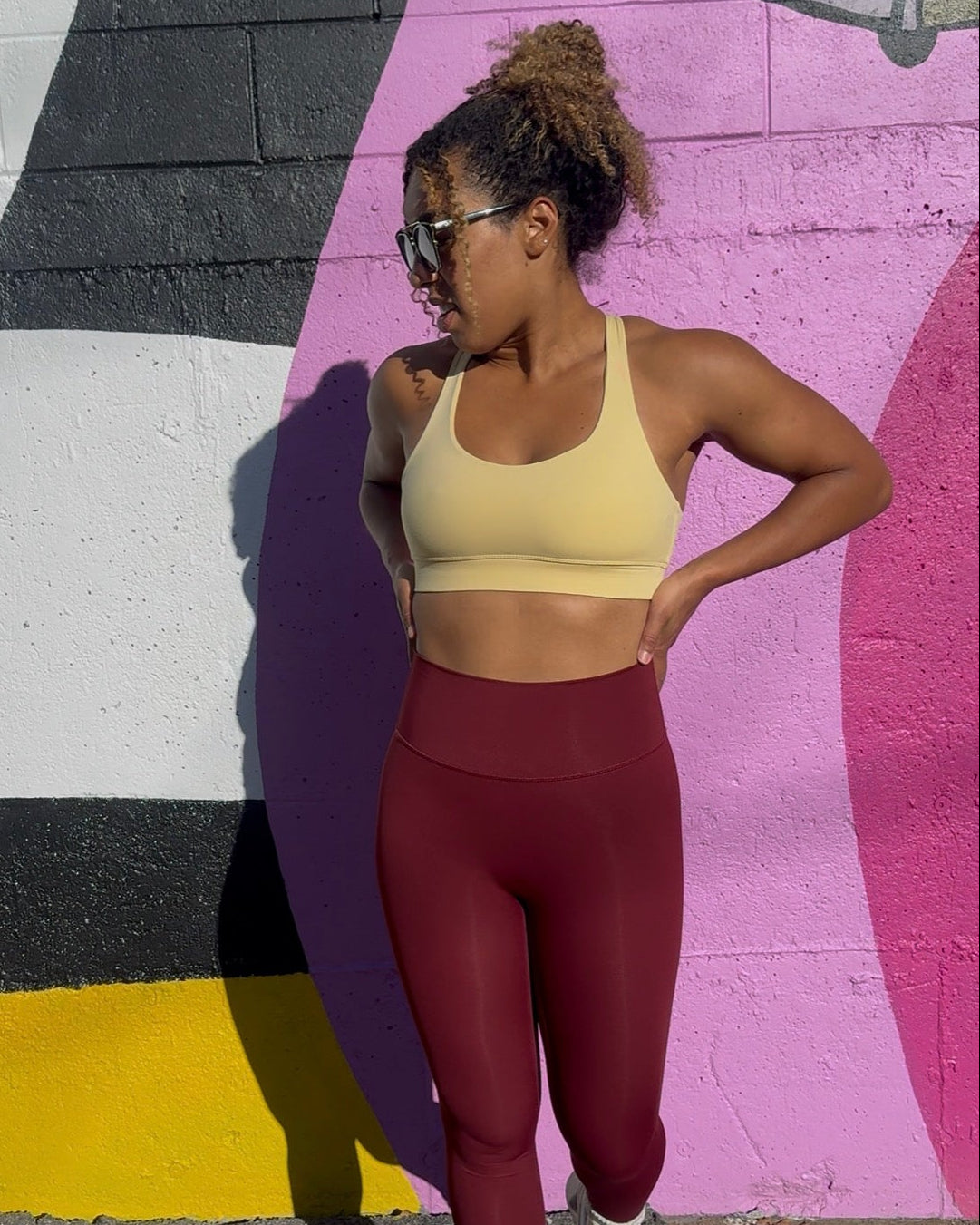 A woman posing outdoors in front of a colorful graffiti wall, wearing a light yellow sports bra and high-waisted burgundy leggings with white sneakers.