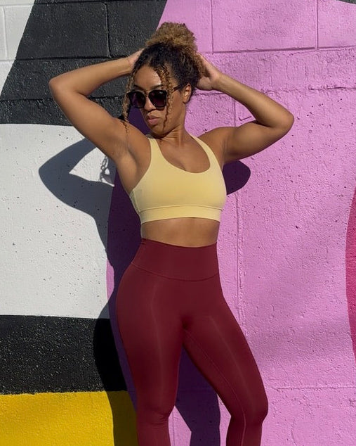 A woman posing outdoors in front of a colorful graffiti wall, wearing a light yellow sports bra and high-waisted burgundy leggings with white sneakers.