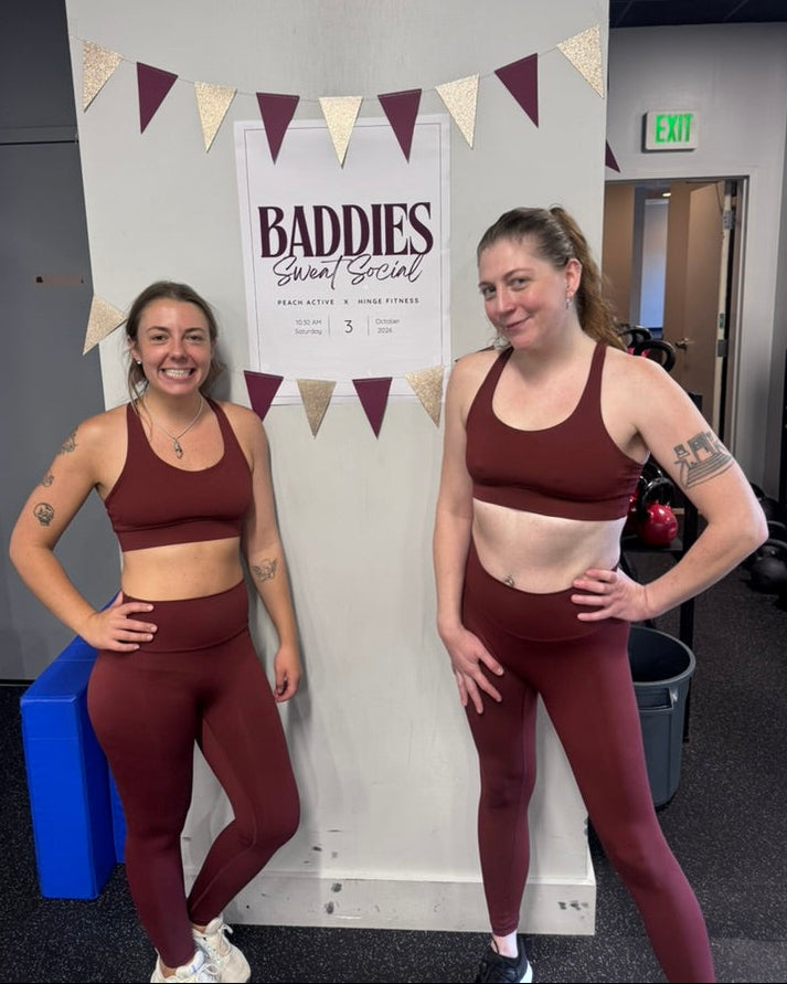Two women posing in matching burgundy sports bra and leggings activewear sets in front of a white wall with a 'Baddies Smart Social' sign and bunting.
