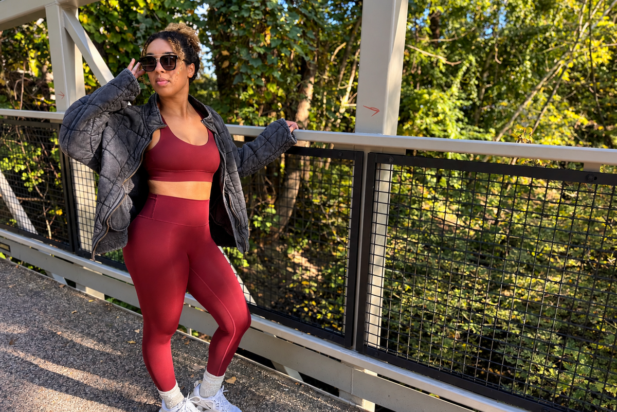 Woman wearing maroon sports bra and leggings, black jacket, sunglasses, standing on a bridge
