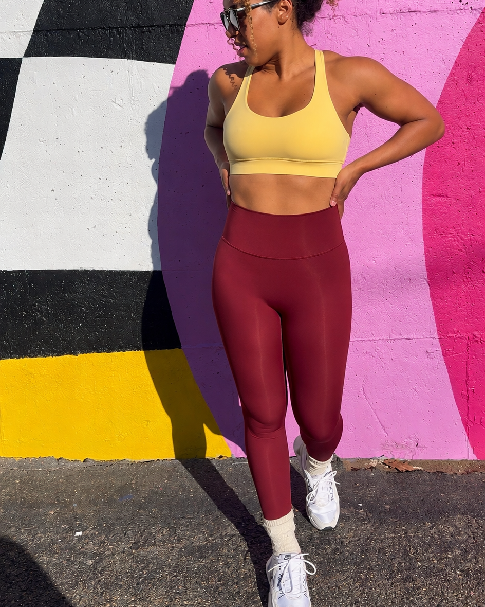 A woman posing outdoors in front of a colorful graffiti wall, wearing a yellow sports bra and high-waisted burgundy leggings with white sneakers.