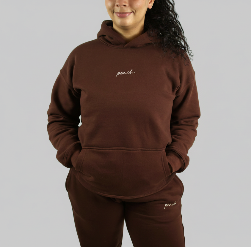 Person wearing a brown hoodie and pants with 'peach' embroidery on a white background