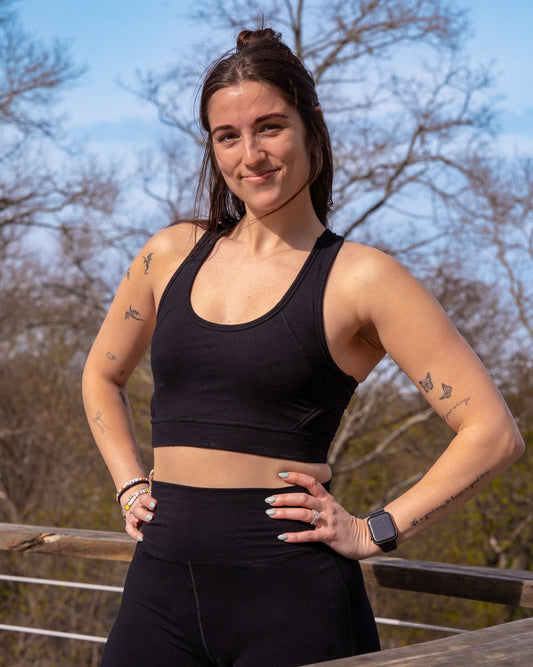 A woman wearing a black sports bra and matching black leggings stands outdoors with her hands on her hips, smiling at the camera against a background of trees and blue sky.