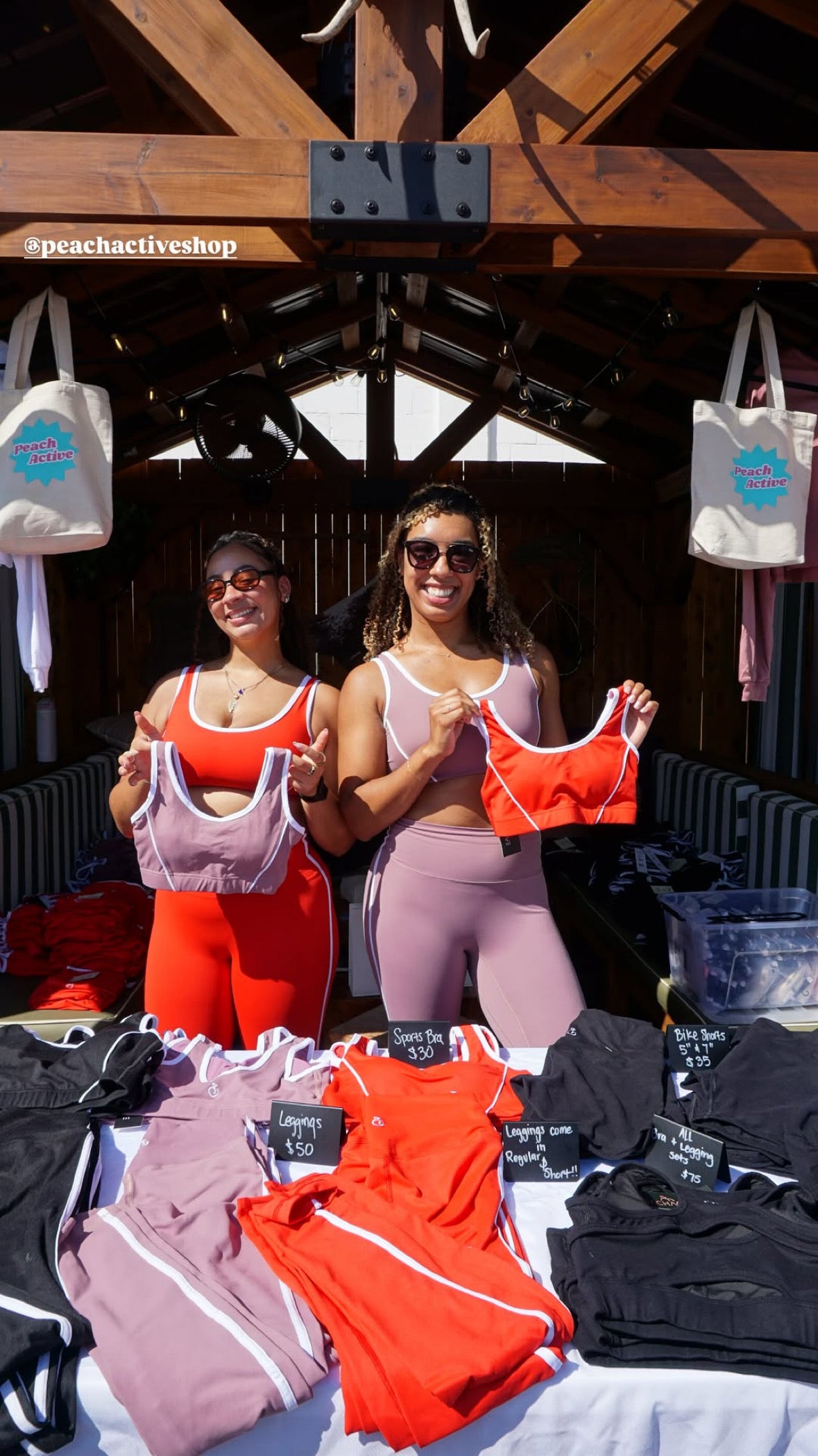 Two women showcasing athletic wear at an outdoor event with branded bags and clothing in the background.