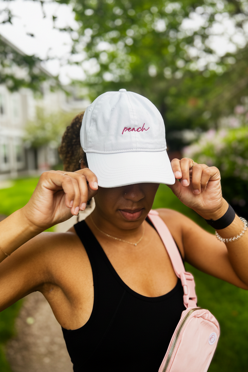 Person wearing a light gray cap with 'peach' embroidered on it, standing outdoors.