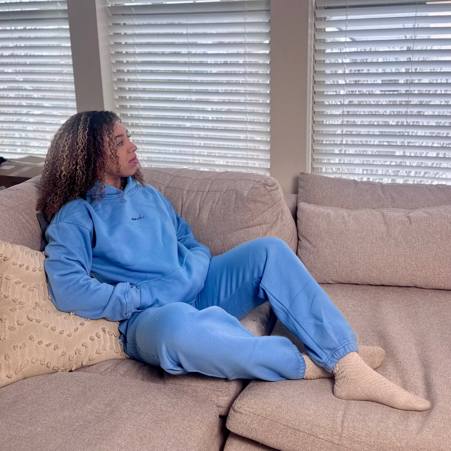 Person wearing a blue outfit sitting on a beige couch with blinds in the background