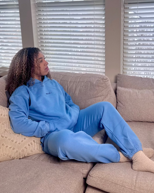 Person wearing a blue outfit sitting on a beige couch with blinds in the background