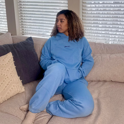 Person wearing a blue tracksuit sitting on a beige couch with blinds in the background