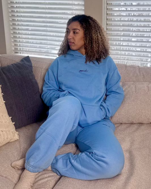 Person wearing a blue tracksuit sitting on a beige couch with blinds in the background