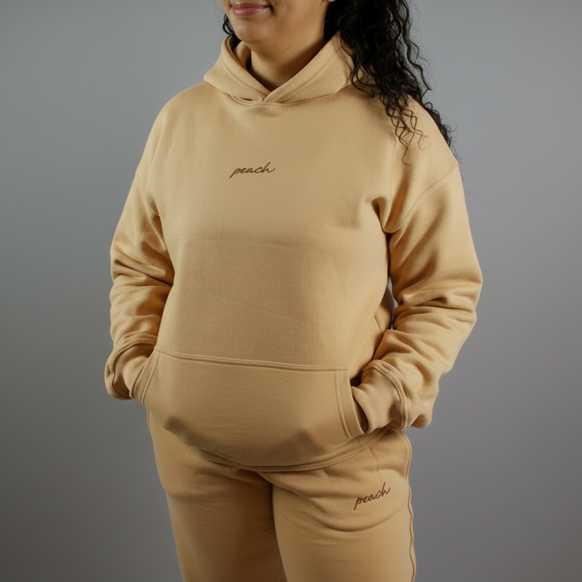 Person wearing a beige hoodie and pants set with 'peach' embroidery on a plain background